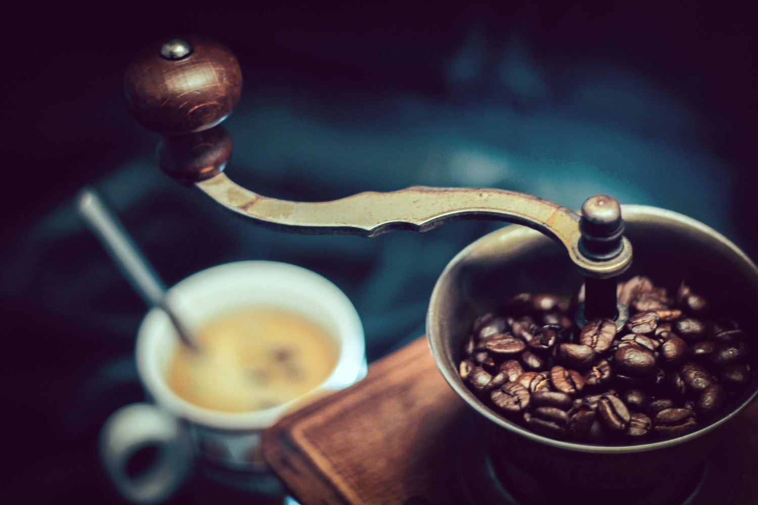 A brass hand grinder full of roasted beans beside a fresh cup of coffee